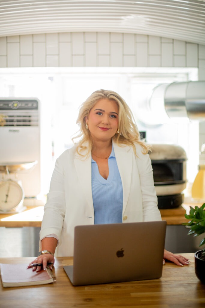 A blonde woman in a white blazer and blue top stands behind a wooden counter with a laptop and a notebook.