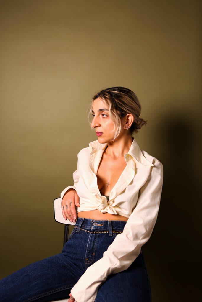 Woman in a cream shirt and jeans seated on a stool against an olive studio backdrop, looking to the side