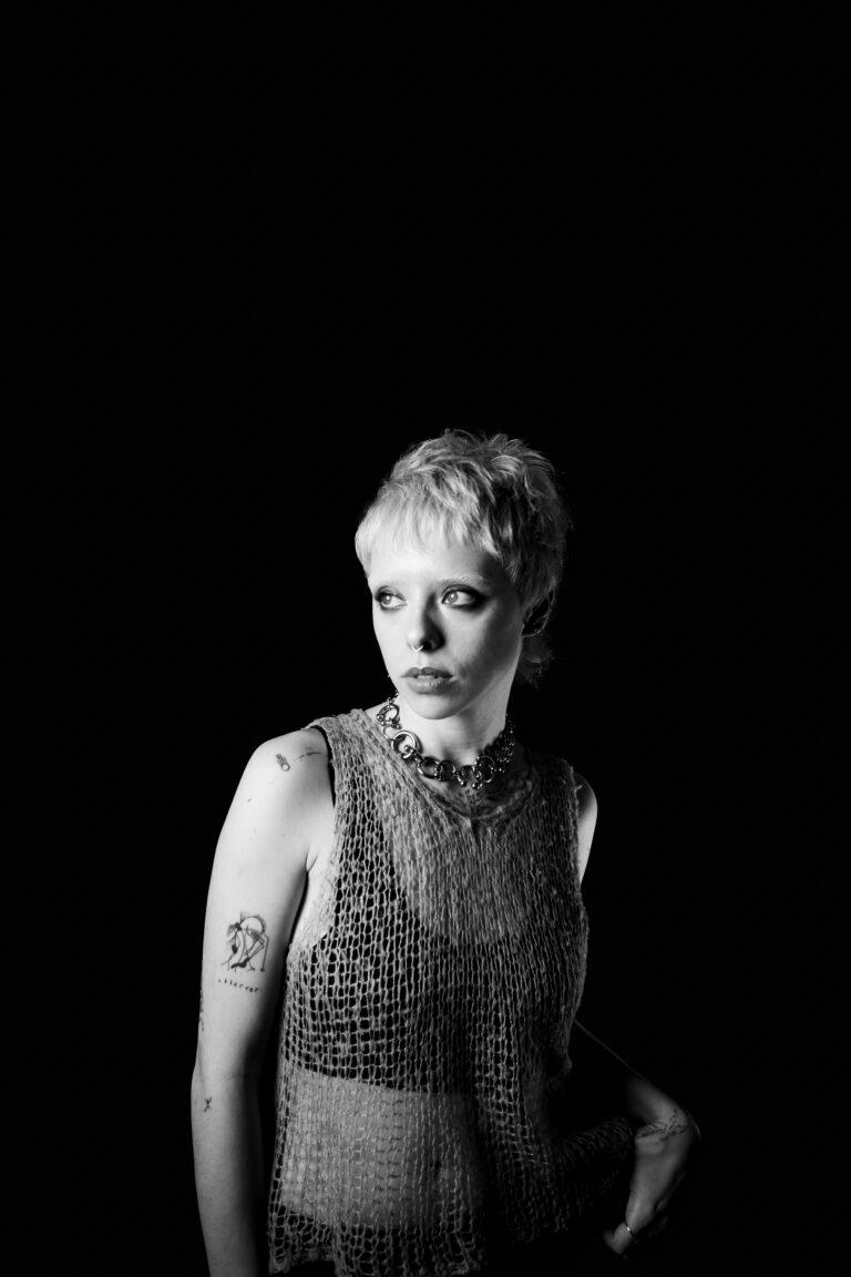 Black and white photography Model Tabby looking at the camera with a sleevely large knit top, large silver necklave and short pixie cut hair with a black background