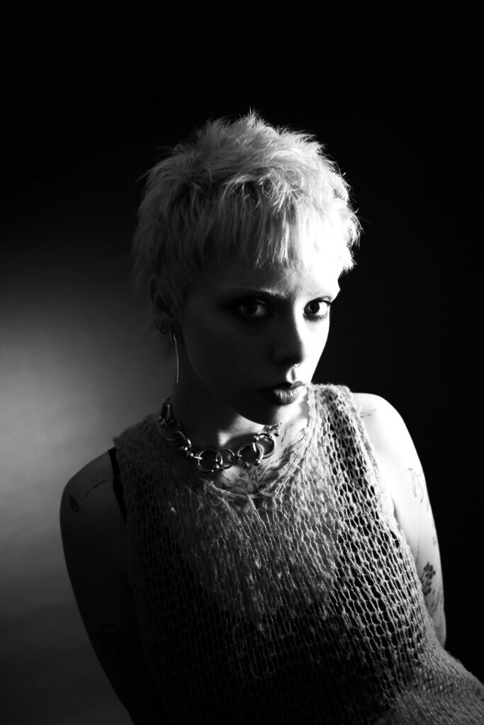 Black and white portrait of a person with short, fair hair, wearing a mesh top and a chunky chain necklace, looking towards the camera with dramatic lighting.
