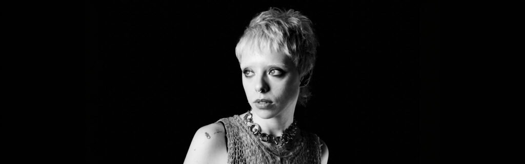 Black and white portrait of a person with short bleached hair and a chunky silver necklace against a black background