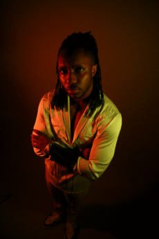 A striking PORTRAIT of a man with long plaited hair stands with arms folded in dramatic lighting, wearing a cream shirt and dark trousers, looking up at the camera—perfect for your BTM-inspired collection.