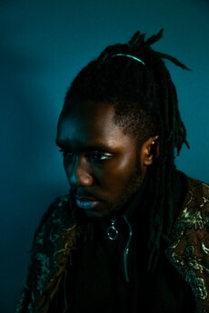 A striking PORTRAIT of a person with dark skin and dreadlocks, wearing a patterned jacket and black top, looking off to the side against a dark blue background.
