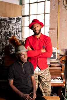 Two men pose in a cluttered room with a piano and a table football table; one sits wearing a hat, the other stands behind him in a red hat and shirt with camouflage trousers.
