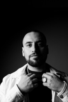 Black and white portrait of DJ Rami Ali with a beard wearing a light jacket, necklace, and ring, facing the camera with his hands near his collar.