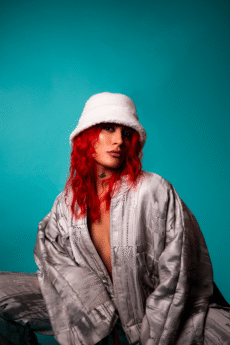 Musician SEVEN with long red hair wears a white bucket hat and silvery patterned jacket, posing against a teal background.
