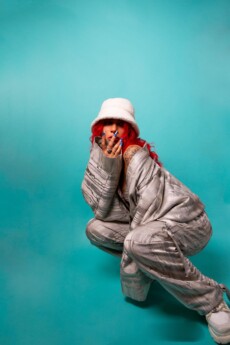 Musician SEVEN with long red hair wears a white bucket hat and silvery patterned outfit, posing against a teal background.