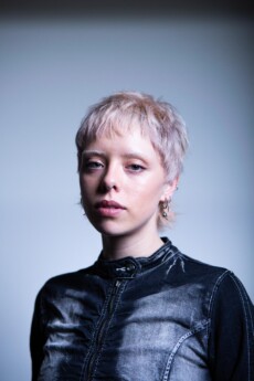 Model Tabby porfolio photograph standing wearing a denim long sleeve top with light blonde hair in a pixie cute in front of a gradient grey background